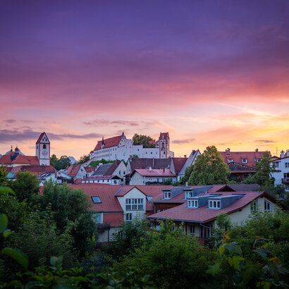  Das Bild fängt eine friedliche Sonnenuntergangsszene in Füssen ein. Der Himmel, der in Farbtönen von Lila und Pink gemalt ist, dient als schöne Kulisse für die Altstadt. Das Bild selbst ist ein charmanter Mix aus weißen und roten Gebäuden, deren Dächer eine Farbtupfer in die Szene bringen. Die allgemeine Atmosphäre des Bildes ist friedvoll und idyllisch und fängt das Wesen eines ruhigen kleinen Ortes in der Abenddämmerung ein.