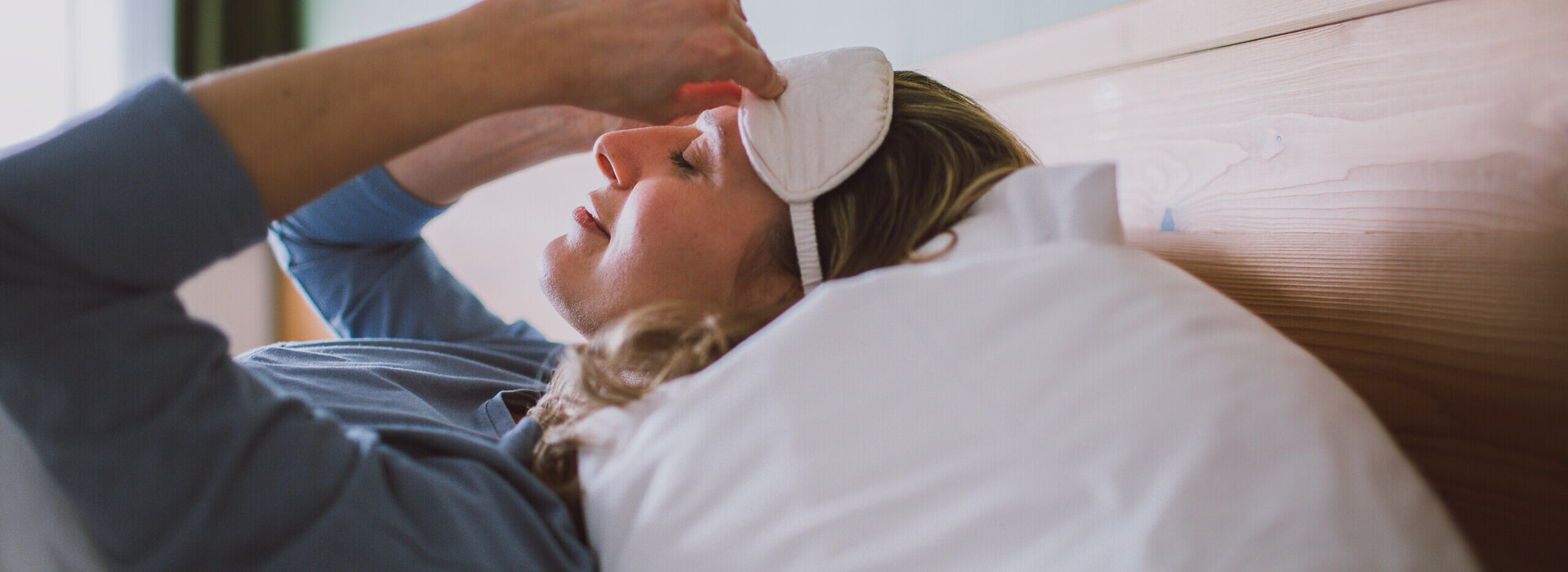 Das Bild zeigt eine Frau mit einem blauen langen Oberteil. Sie liegt in einem Bett, das weiß bezogen ist und zieht sich gerade eine weiße Schlafmaske an.