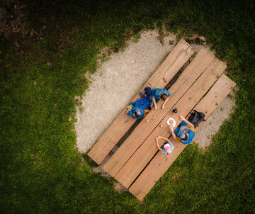 Das Bild zeigt eine Familie, bestehend aus einer Frau einem Mann, einem Mädchen und einem Jungen. Sie tragen sportliche Freizeitkleidung. Sie sitzen an einem Tisch mit zwei Holzbänken und essen. Das Foto ist aus der Vogelperspektive aufgenommen.  