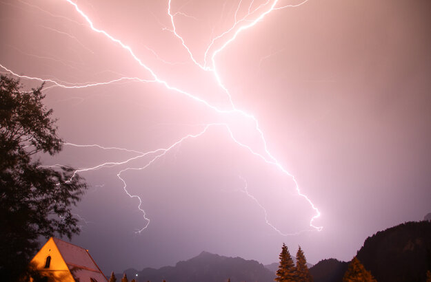 Das Bild zeigt einen weit verästelten und hell erleuchteten Gewitterblitz am Himmel. Der Himmel drum herum ist in dunkles lila getaucht. Im unteren Bereich des Bildes sieht man Berge, bewaldete Hügel und Wälder. Sowie ein beleuchtetes Hausdach. 