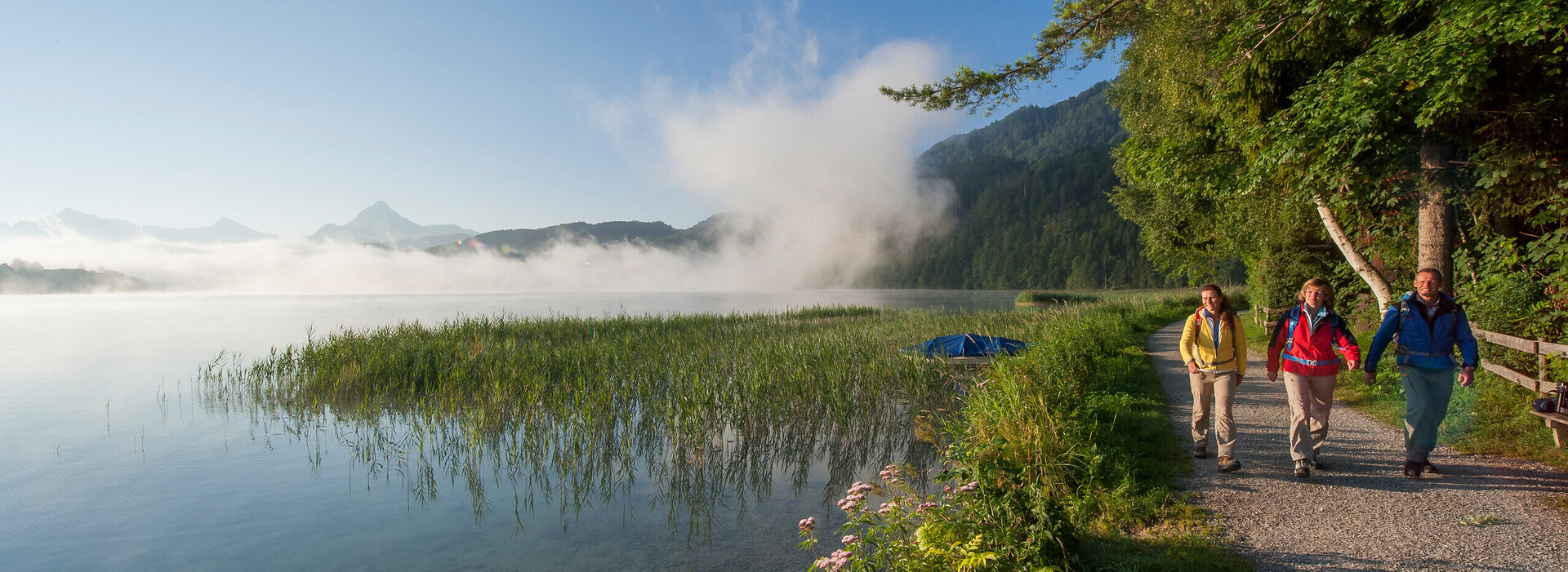 Das Bild zeigt ein sonniges Seeufer mit Bergen die im Nebel liegen. Am Ufer entlang wandern zwei Frauen und ein Mann.