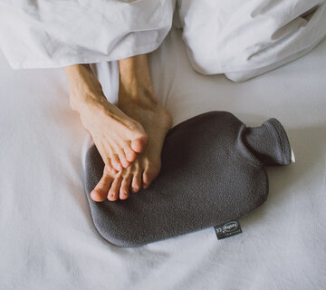 Das Bild zeigt zwei Füße, die auf einer grauen Wärmflasche liegen und unter einer weißen Bettdecke hervorspitzeln.  