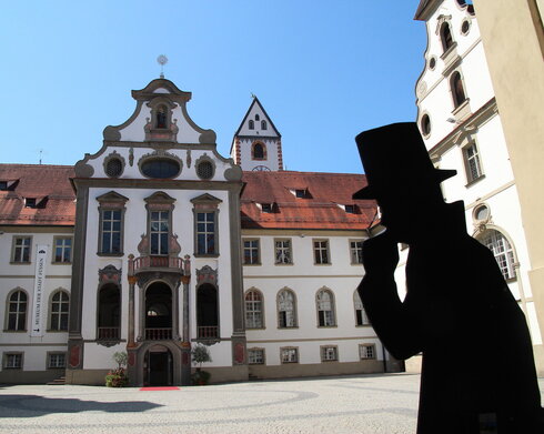 Das Foto zeigt den Klosterhof des ehemaligen Benediktinerklosters St. Mang. Der Blick fällt auf den Eingang des Museums der Stadt Füssen. Der Boden des Hofs ist mit Kopfsteinpflaster gepflastert. Die Fassade des Gebäudes ist mit Illusionsmalerei verziert. Vor dem Eingang des Museums liegt ein roter Teppich auf dem Boden. Der Himmel ist blau. Die Sonne scheint.