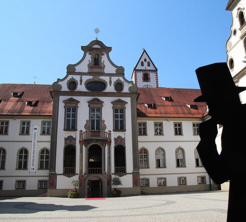 Das Foto zeigt den Klosterhof des ehemaligen Benediktinerklosters St. Mang. Der Blick fällt auf den Eingang des Museums der Stadt Füssen. Der Boden des Hofs ist mit Kopfsteinpflaster gepflastert. Die Fassade des Gebäudes ist mit Illusionsmalerei verziert. Vor dem Eingang des Museums liegt ein roter Teppich auf dem Boden. Der Himmel ist blau. Die Sonne scheint.