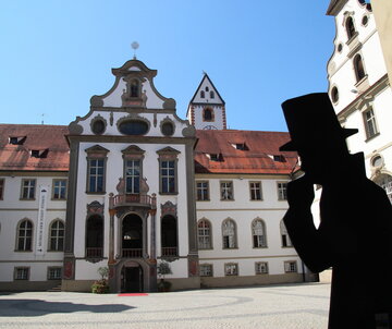 Das Foto zeigt den Klosterhof des ehemaligen Benediktinerklosters St. Mang. Der Blick fällt auf den Eingang des Museums der Stadt Füssen. Der Boden des Hofs ist mit Kopfsteinpflaster gepflastert. Die Fassade des Gebäudes ist mit Illusionsmalerei verziert. Vor dem Eingang des Museums liegt ein roter Teppich auf dem Boden. Der Himmel ist blau. Die Sonne scheint.