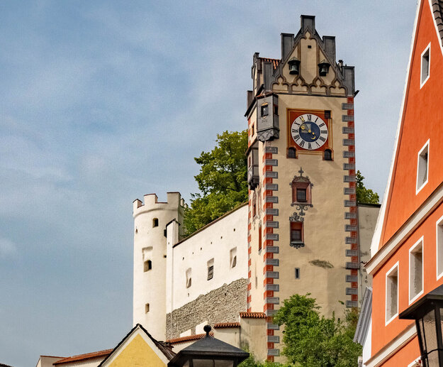 Das Foto zeigt den Uhrturm des Hohen Schlosses und den an den Turm angrenzenden Gebäudeteil. Darunter sieht man die bunten Fassaden von Altstadtgebäuden. Die Straßenlaternen sind mit Blumenkästen geschmückt, in denen Geranien und andere Blumen blühen. Die Sonne scheint.