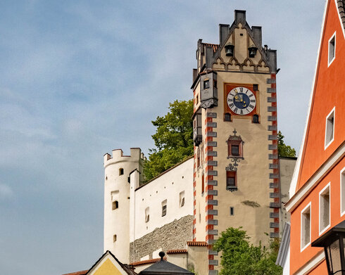 Das Foto zeigt den Uhrturm des Hohen Schlosses und den an den Turm angrenzenden Gebäudeteil. Darunter sieht man die bunten Fassaden von Altstadtgebäuden. Die Straßenlaternen sind mit Blumenkästen geschmückt, in denen Geranien und andere Blumen blühen. Die Sonne scheint. 