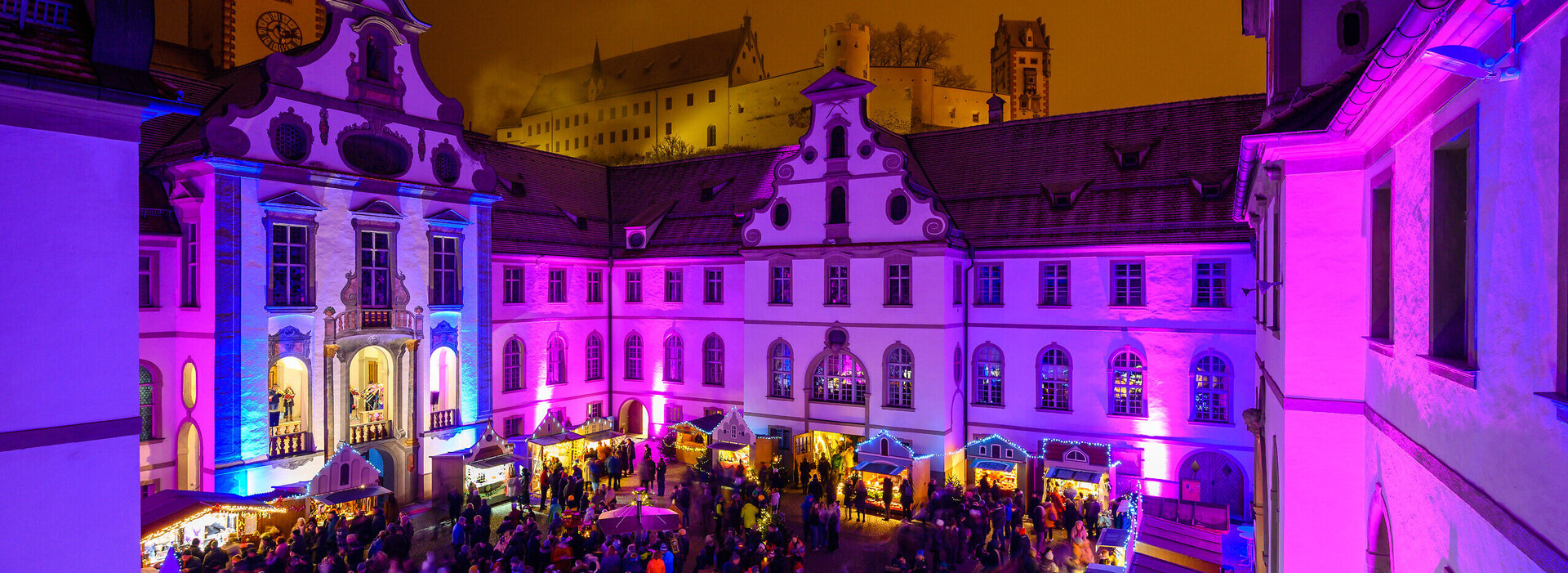 Das Bild fängt eine lebendige nächtliche Szene in Füssen auf dem Adventsmarkt ein. Die Skyline wird dominiert von einem großen, verzierten Gebäude mit Uhrturm, das sich deutlich gegen den Nachthimmel abhebt. Das Gebäude ist mit lila und pinken Lichtern beleuchtet, was eine fröhliche Atmosphäre schafft. Die Straße vor dem Gebäude ist von Menschen belebt, die ebenfalls von den gleichen lila und pinken Lichtern erhellt werden. Die Menge scheint ein Fest oder eine Veranstaltung zu genießen, wie die bunten Lichter und die lebendige Atmosphäre darauf hindeuten. Die Gesamtansicht vermittelt das Gefühl von Feier und Gemeinschaftsgeist.