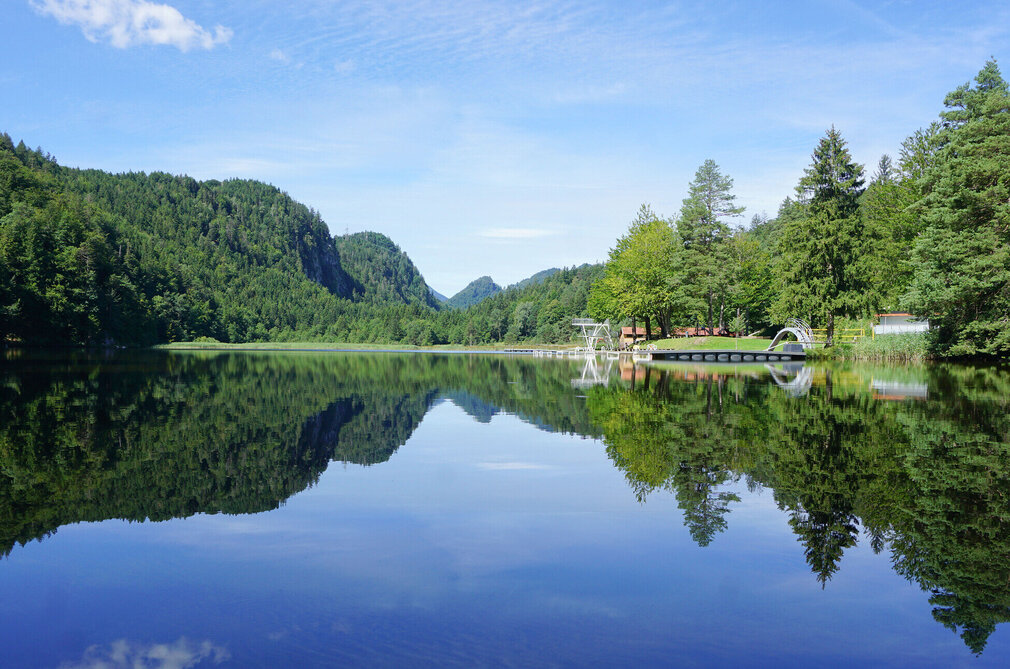 Das Bild zeigt eine ruhige Seenlandschaft. Der Obersee, der der zentrale Fokus des Bildes ist, ist von üppigem Grün umgeben und von Bergen gesäumt. Die ruhige Wasseroberfläche des Sees spiegelt die Umgebung wider. Auf der rechten Seite des Bildes befindet sich ein Steg mit einem Sprungturm. Der Himmel darüber ist klar und blau.
