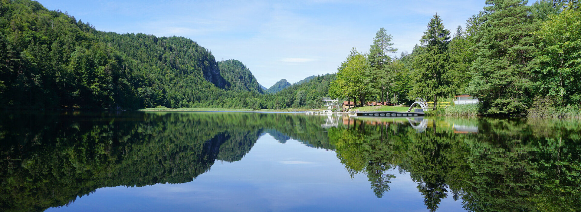 Das Bild zeigt eine ruhige Seenlandschaft. Der Obersee, der der zentrale Fokus des Bildes ist, ist von üppigem Grün umgeben und von Bergen gesäumt. Die ruhige Wasseroberfläche des Sees spiegelt die Umgebung wider. Auf der rechten Seite des Bildes befindet sich ein Steg mit einem Sprungturm. Der Himmel darüber ist klar und blau.