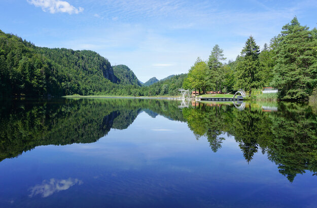 Das Bild zeigt eine ruhige Seenlandschaft. Der Obersee, der der zentrale Fokus des Bildes ist, ist von üppigem Grün umgeben und von Bergen gesäumt. Die ruhige Wasseroberfläche des Sees spiegelt die Umgebung wider. Auf der rechten Seite des Bildes befindet sich ein Steg mit einem Sprungturm. Der Himmel darüber ist klar und blau.