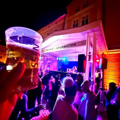 Das Bild zeigt eine Hausfassade. Unter dem überdachten Haupteingang steht ein beleuchtets DJ-Pult. Darüber hängt ein Schild Tourist Information. Viele Leute tanzen. Sie tragen Mützen und Winterkleidung. Im Vordergrund hält eine Hand einen Becher mit Bier gefüllt. Auf dem Becher steht „Füssen“.  