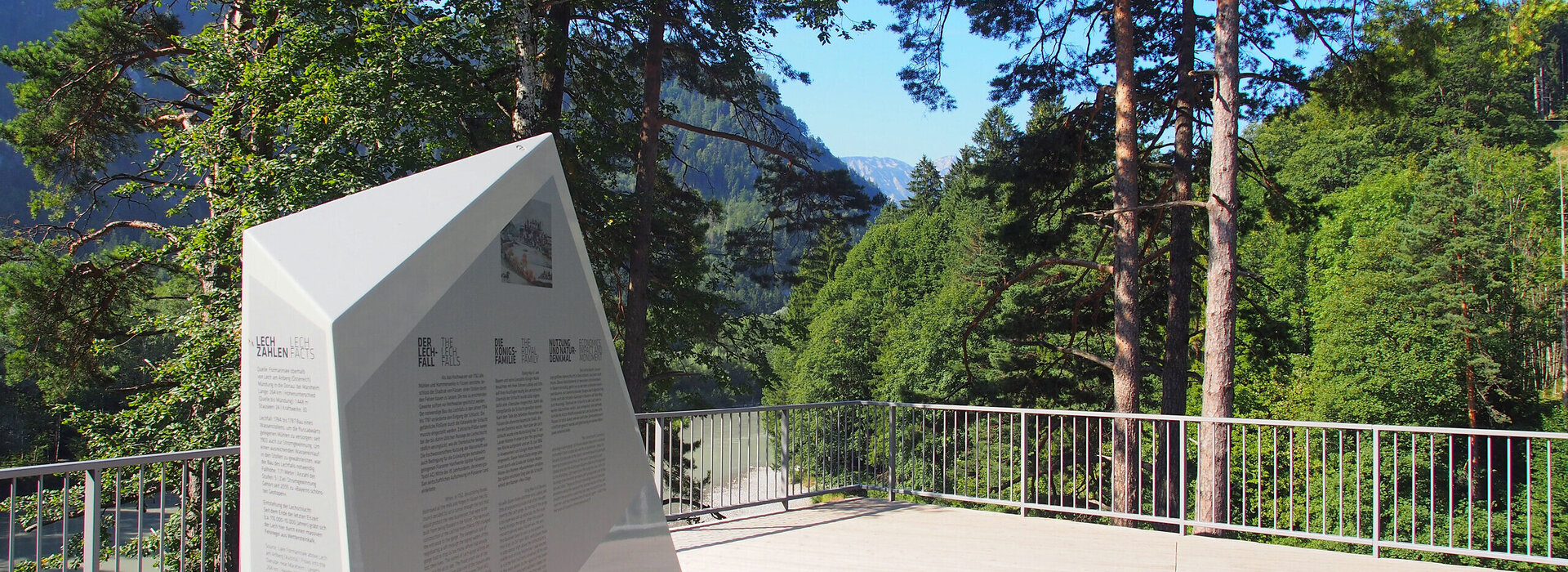 Das Bild zeigt eine idyllische Außenlandschaft, die ein deutlich sichtbar angebrachtes, großes, weißes und dreieckiges Schild auf einem Holzdeck umfasst. Das Schild ist mit schwarzer Schrift verziert, der Inhalt des Textes ist jedoch in dem Bild nicht ersichtlich. Das Deck, das aus Holzbohlen erbaut ist, befindet sich auf einem Hügel und bietet einen Ausblick auf Bäume und einen Bergkamm in der Ferne. Das Deck wird von einem Metallgeländer umgeben, was Sicherheit beim Genießen der Aussicht gewährleistet. Die Szene transportiert insgesamt ein Gefühl von Ruhe und natürlicher Schönheit.
