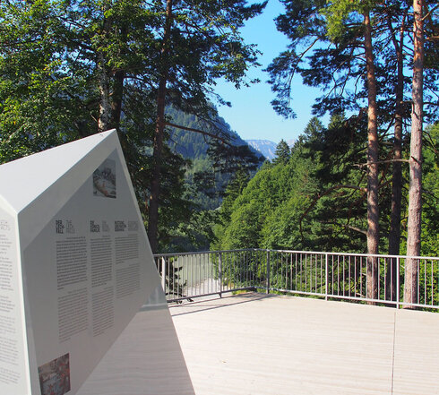 Das Bild zeigt eine idyllische Außenlandschaft, die ein deutlich sichtbar angebrachtes, großes, weißes und dreieckiges Schild auf einem Holzdeck umfasst. Das Schild ist mit schwarzer Schrift verziert, der Inhalt des Textes ist jedoch in dem Bild nicht ersichtlich. Das Deck, das aus Holzbohlen erbaut ist, befindet sich auf einem Hügel und bietet einen Ausblick auf Bäume und einen Bergkamm in der Ferne. Das Deck wird von einem Metallgeländer umgeben, was Sicherheit beim Genießen der Aussicht gewährleistet. Die Szene transportiert insgesamt ein Gefühl von Ruhe und natürlicher Schönheit.