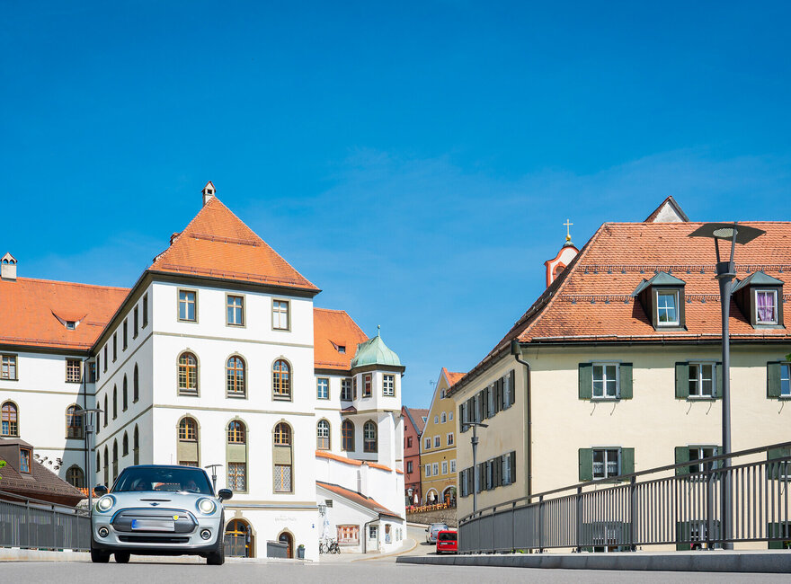 Ein silberner Mini fährt auf den Betrachter zu. Er fährt über eine Brücke. Im Hintergrund erhebt sich neben den bunten Altstadthäusern, der Gebäudekomplex des ehemaligen Benediktinerklosters mit seinen roten Dächern und unzähligen Fenstern. Der Himmel ist blau. Die Sonne scheint. 