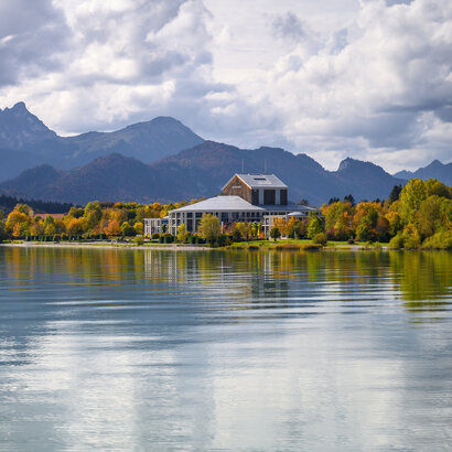 Das Foto wurde vom See aus aufgenommen. Am Ufer steht ein prächtiges Gebäude mit einem grauen Dach und vielen Fenstern. Der Mittelteil des Gebäudes ist rund. Es handelt sich um das Festspielhaus Neuschwanstein. Die Bäume ringsum haben schon eine bunte Farbe angenommen. Im Hintergrund erstreckt sich die Alpenkette. Der Himmel ist wolkenverhangen, trotzdem scheint die Sonne. 