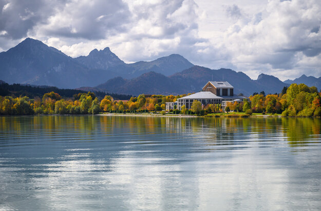 Das Foto wurde vom See aus aufgenommen. Am Ufer steht ein prächtiges Gebäude mit einem grauen Dach und vielen Fenstern. Der Mittelteil des Gebäudes ist rund. Es handelt sich um das Festspielhaus Neuschwanstein. Die Bäume ringsum haben schon eine bunte Farbe angenommen. Im Hintergrund erstreckt sich die Alpenkette. Der Himmel ist wolkenverhangen, trotzdem scheint die Sonne. 