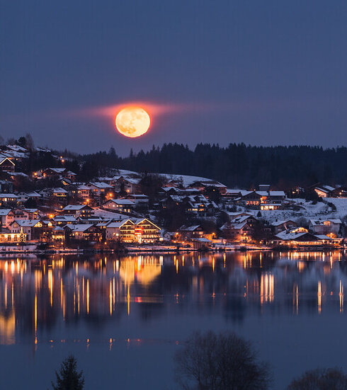 Das Bild fängt eine ruhige, winterliche Nachtszene im Ort Hopfen ein, der am Ufer des Hopfensees liegt. Der Ort wird von dem weichen Licht der Straßen- und Häuserbeleuchtung in warmes Licht getaucht, das sich im ruhigen Wasser des Sees spiegelt. In der Ferne erheben sich majestätische, schneebedeckte Berge gegen den dunklen Himmel. Der Nachthimmel wird vom Vollmond erhellt, der über die ganze Szene strahlt. 