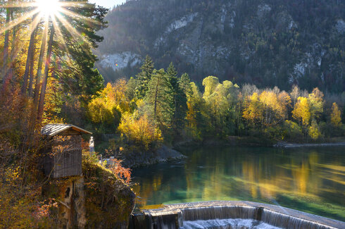 Das Bild zeigt den Lechfall bei Füssen. Hier wir der Fluss Lech über stufen nach unten geleitet. Das Bachbett ist von hohen Bäume gesäumt, durch deren Äste und Zweige die Sonne scheint. Im Hintergrund erheben sich bewaldete Hügel. Der Himmel ist blau. 
