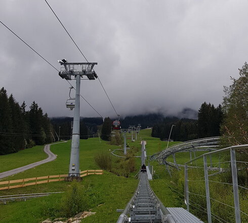 Das Bild zeigt auf der linken Bildseite eine Sesselbahn. Auf der rechten Seite ein Eisengestell, dass einen Schlitten nach oben zieht. Der Hang ist mit grünem Gras bewachsen. Der Himmel ist wolkenverhangen.   
