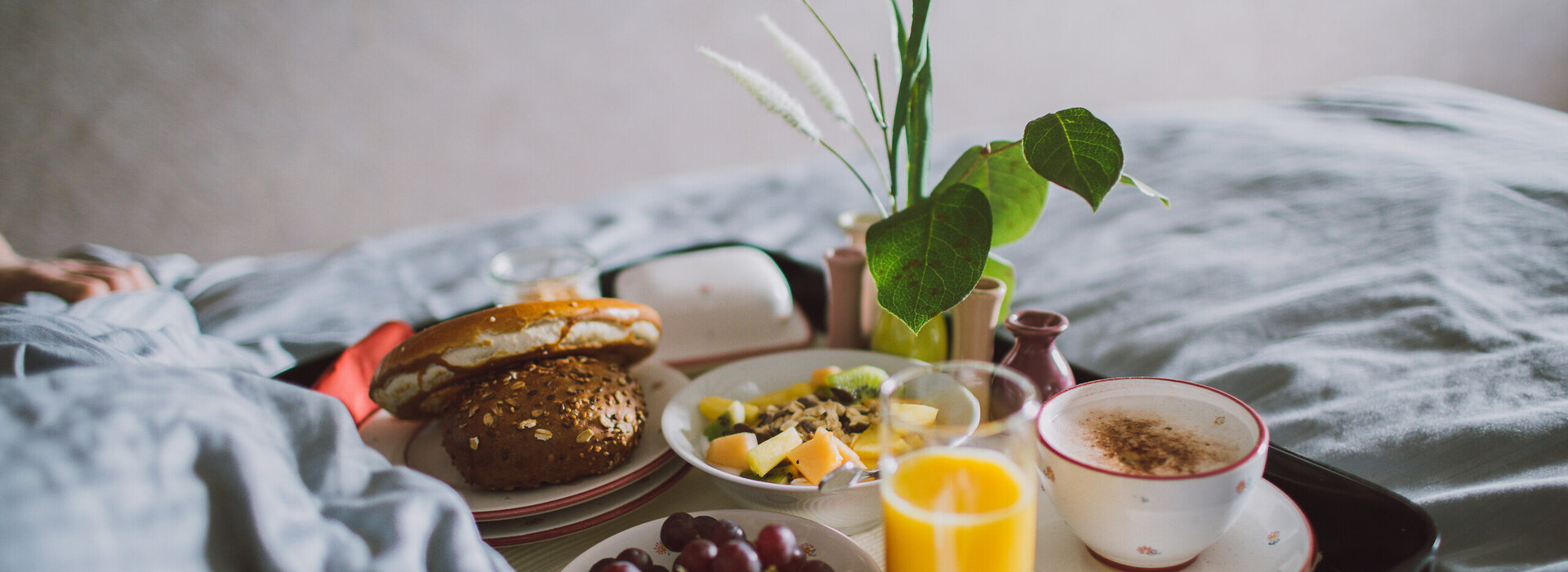 Das Bild zeigt ein Frühstückstablett, auf dem zwei Tellern, Tassen, Müsli, Butter, einem Kännchen stehen. Das Tablett steh auf einer grauen Bettdecke. 