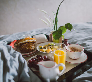 Das Bild zeigt ein Frühstückstablett, auf dem zwei Tellern, Tassen, Müsli, Butter, einem Kännchen stehen. Das Tablett steh auf einer grauen Bettdecke. 