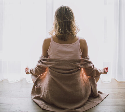 Das Bild zeigt eine Frau, die im Schneidersitz auf dem Holzboden sitzt. Sie sitz vor einem hellen Fenster, das mit einem bodenlangen Stohr verhängt ist. Sie eine beige Strickjacke, die ihr von den Schultern gerutscht ist, ein rosafarbenes Tanktop und eine kurze Hose. Ihre Hände ruhen auf ihren Knien. Mittelfinger und Daumen beider Hände berühren sich. Sie hat dem Betrachter den Rücken zugedreht und ihren Blick auf das helle Fenster gerichtet.    