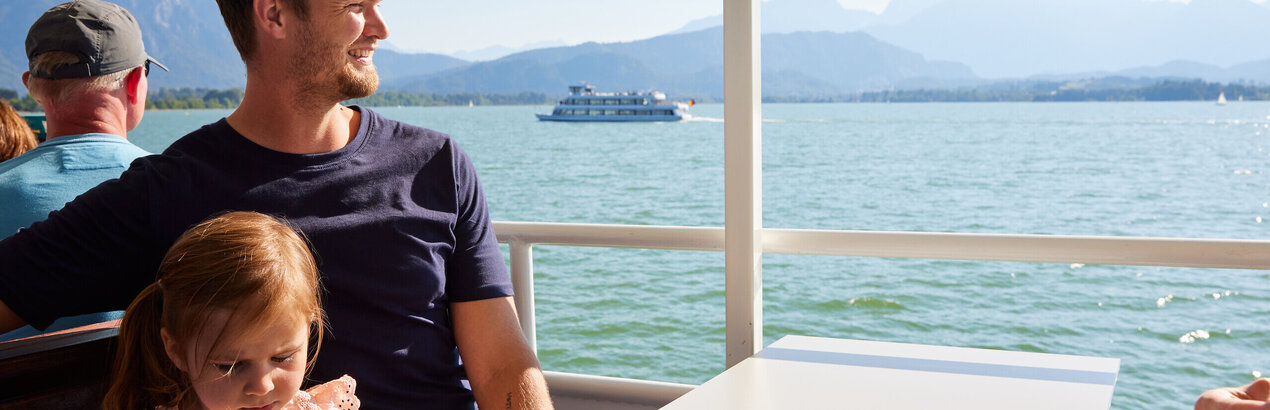 Das Bild fängt eine ruhige Szene auf einem Boot ein, wo ein Mann und ein junges Mädchen an einem Tisch sitzen und ein Essen gemeinsam genießen. Der Mann, der ein blaues Hemd trägt, sitzt auf der linken Seite des Tisches, während das Mädchen, das ein rosa Hemd trägt, auf der rechten Seite sitzt. Der Tisch ist mit einer weißen Tasse, einer weißen Schüssel und einer weißen Flasche geschmückt, was auf eine legere und entspannte Essenszeit hinweist.  Das Boot, auf dem sie sind, segelt über einem weiten Gewässer, mit majestätischen Bergen, die eine malerische Kulisse bilden. Der klare blaue Himmel über ihnen und das ruhige Wasser unter ihnen tragen zur Ruhe der Szene bei. Die Gesamtatmosphäre des Bildes ist eine von Frieden und Zusammengehörigkeit, wenn der Mann und das Mädchen ein Essen teilen und dabei die atemberaubende Umgebung um sie herum genießen.