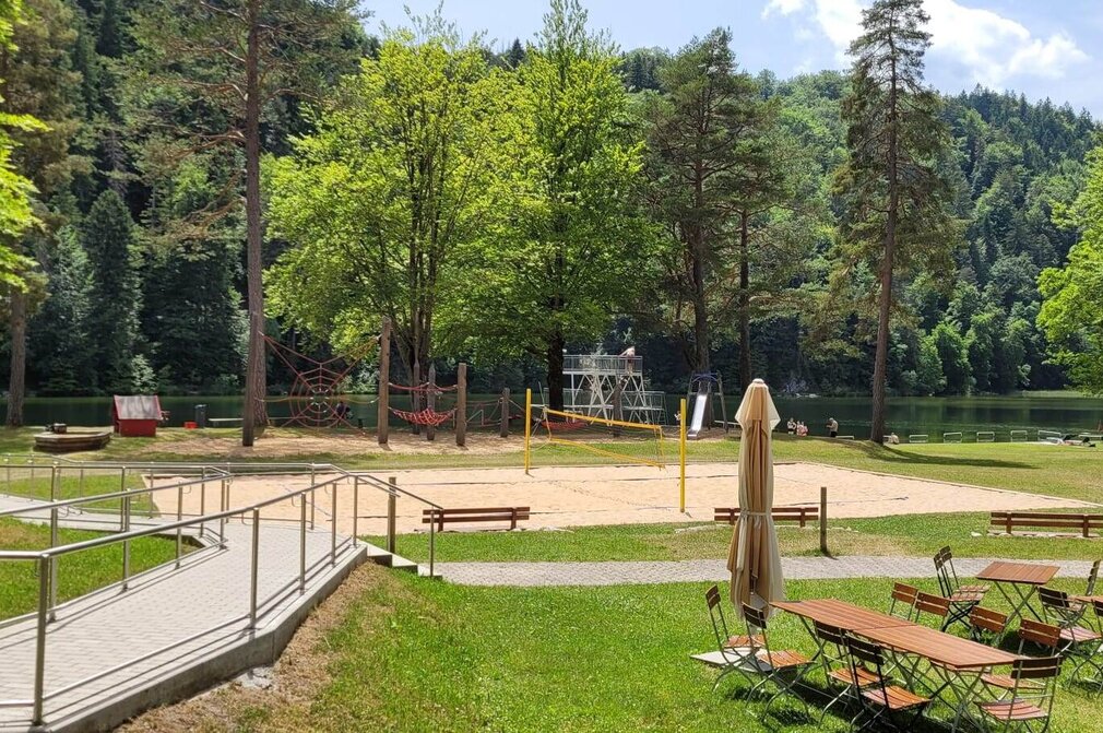 Das Bild zeigt einen Blick ins Gelände des Oberseebads. Das zentrale Motiv ist ein sandiger Volleyballplatz, umgeben von üppigem Grün, Bäumen und Gras. Der Platz ist mit weißen Linien markiert, die die Grenzen für das Spiel anzeigen. Im Vordergrund befinden sich mehrere hölzerne Bänke und Tische, die in einem Halbkreis um den Platz angeordnet sind.  In der Ferne liegt der ruhige Obersee. Die Oberfläche des Wassers ist unberührt und spiegelt die umliegende Vegetation wider. Der Himmel darüber ist klar und blau mit einzelnen, verstreuten Wolken.