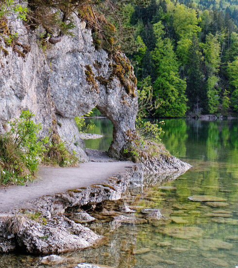 Das Bild zeigt einen See. Im linken Bildrand ist der Uferbereich zu sehen, an dem eine Felswand emporsteigt. Der Felsen bildet ein Art „Tor“, durch das sich der Weg fortsetzt. Der Uferweg vor dem „Felsentor“ ist befestigt. Das Wasser ist klar. Im Hintergrund ragt der Bergwald in die Höhe. Die Bäume sind in unterschiedliche Grüntöne getaucht. Im Vordergrund des Bildes kann man den steinigen Seeboden durch das klare Wasser sehen.   