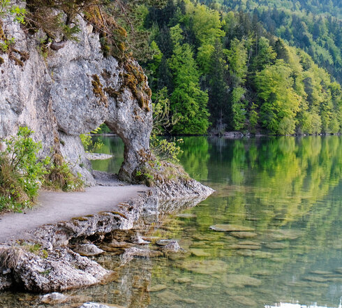 Das Bild zeigt einen See. Im linken Bildrand ist der Uferbereich zu sehen, an dem eine Felswand emporsteigt. Der Felsen bildet ein Art „Tor“, durch das sich der Weg fortsetzt. Der Uferweg vor dem „Felsentor“ ist befestigt. Das Wasser ist klar. Im Hintergrund ragt der Bergwald in die Höhe. Die Bäume sind in unterschiedliche Grüntöne getaucht. Im Vordergrund des Bildes kann man den steinigen Seeboden durch das klare Wasser sehen.   