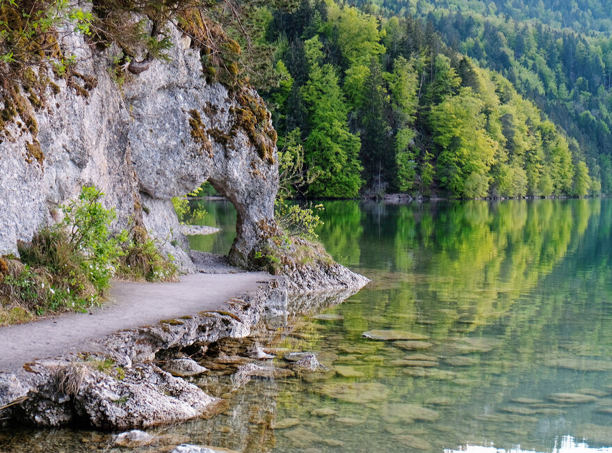 Das Bild zeigt einen See. Im linken Bildrand ist der Uferbereich zu sehen, an dem eine Felswand emporsteigt. Der Felsen bildet ein Art „Tor“, durch das sich der Weg fortsetzt. Der Uferweg vor dem „Felsentor“ ist befestigt. Das Wasser ist klar. Im Hintergrund ragt der Bergwald in die Höhe. Die Bäume sind in unterschiedliche Grüntöne getaucht. Im Vordergrund des Bildes kann man den steinigen Seeboden durch das klare Wasser sehen.   