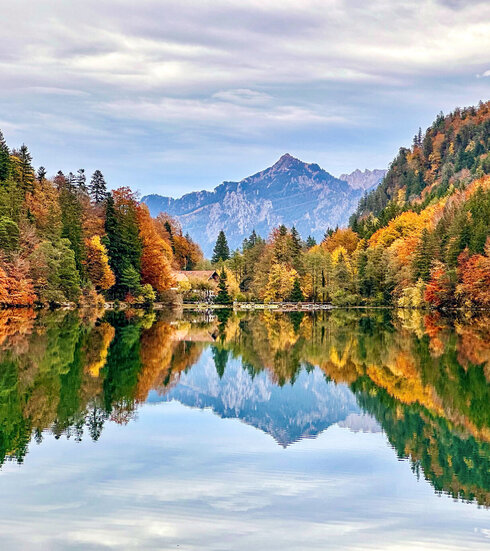 Im Fokus des Bildes liegt ein See, dessen Oberfläche glatt wie ein Spiegel, die umliegende Natur widerspiegelt. Den See säumen bunte Wälder. Im Hintergrund erheben sich die Berge.  