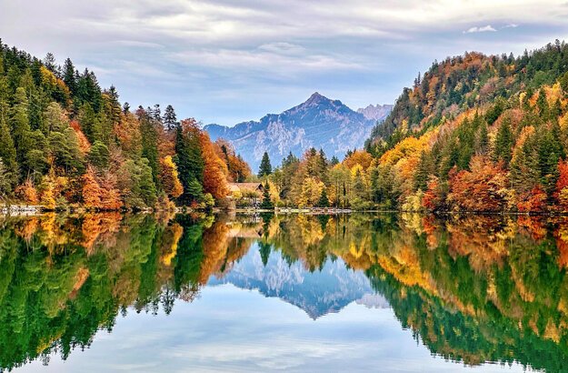 Im Fokus des Bildes liegt ein See, dessen Oberfläche glatt wie ein Spiegel, die umliegende Natur widerspiegelt. Den See säumen bunte Wälder. Im Hintergrund erheben sich die Berge.  