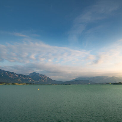 Das Bild zeigt eine idyllische Landschaft, die von einem weiten Gewässer dominiert wird, das sich zum Horizont hin erstreckt – dem Forggensee. Der türkise See ist ruhig und ungestört und spiegelt den klaren Himmel darüber wider. Der Himmel ist mit einigen verstreuten Wolken durchsetzt, die der Szene Tiefe und Dimension verleihen. In der Ferne erheben sich majestätische Berge, deren Gipfel den Himmel berühren. Die Berge, mit ihrem rauen Gelände, bieten einen scharfen Kontrast zum ruhigen Wasser. 