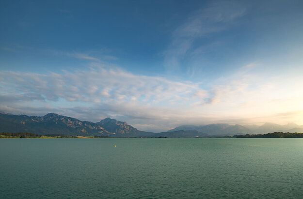 Das Bild zeigt eine idyllische Landschaft, die von einem weiten Gewässer dominiert wird, das sich zum Horizont hin erstreckt – dem Forggensee. Der türkise See ist ruhig und ungestört und spiegelt den klaren Himmel darüber wider. Der Himmel ist mit einigen verstreuten Wolken durchsetzt, die der Szene Tiefe und Dimension verleihen. In der Ferne erheben sich majestätische Berge, deren Gipfel den Himmel berühren. Die Berge, mit ihrem rauen Gelände, bieten einen scharfen Kontrast zum ruhigen Wasser. 