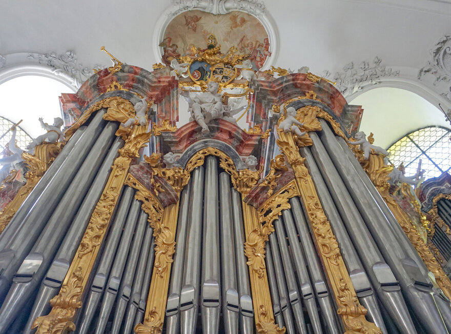 Das Bild zeigt die Pfeifen einer Kirchenorgel, die mit buntem Stuck ringsum reich verziert sind. Das Foto ist von unten aufgenommen.  