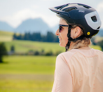 Das Bild zeigt eine Frau auf einem Fahrrad. Sie hält im Moment des Fotos inne und schaut sich um. Dabei trägt sie einen schwarz-weißen Helm mit dem Wort "POC" darauf. Zusätzlich trägt sie eine Sonnenbrille und ein pinkfarbenes Oberteil. Sie hält an den Lenkergriffen ihres Fahrrades fest. Das Feld, auf dem sie fährt, ist grün und reich an Vegetation, mit Bergen im Hintergrund. Der Himmel über ihr ist blau, was auf einen klaren und sonnigen Tag hinweist. Die Frau scheint ihre Fahrt zu genießen und nimmt die schöne Landschaft um sie herum wahr.
