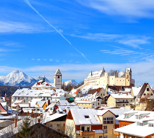 Das Foto zeigt die verschneiten Dächer von Füssen. Über den Dächern thront auf deiner Anhöhe das Hohe Schloss, daneben die St. Mang Klosterkirche. Im Hintergrund sind die verschneiten Berge zu sehen. Der Himmel ist blau. Die Sonne scheint. Es ist Winter.