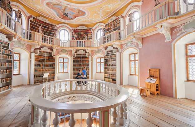 Das Bild zeigt eine imposante, kreisrunde Bibliothek mit einer hohen Decke. Die Wände sind in einem warmen, rosafarbenen Ton gemalt, und die Decke ist mit aufwendigen Mustern und wunderschönen Verzierungen geschmückt.