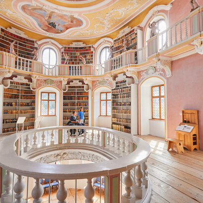 Das Bild zeigt eine imposante, kreisrunde Bibliothek mit einer hohen Decke. Die Wände sind in einem warmen, rosafarbenen Ton gemalt, und die Decke ist mit aufwendigen Mustern und wunderschönen Verzierungen geschmückt.