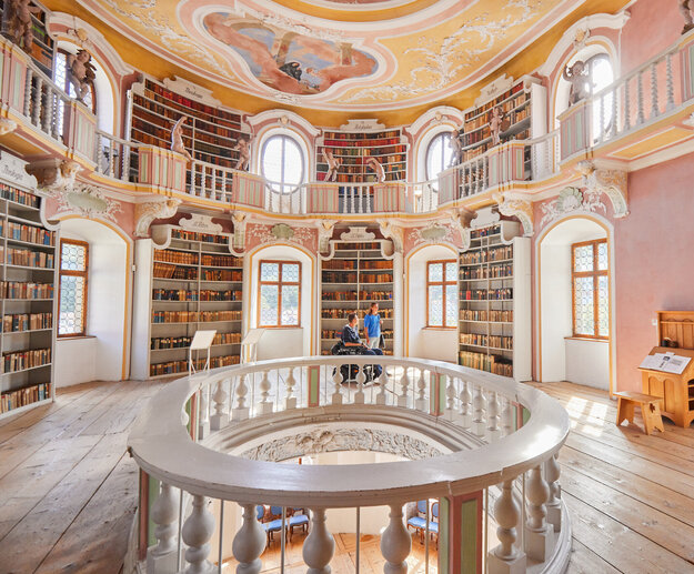 Ehemalige Klosterbibliothek Das Bild zeigt eine imposante, kreisrunde Bibliothek mit einer hohen Decke. Die Wände sind in einem warmen, rosafarbenen Ton gemalt, und die Decke ist mit aufwendigen Mustern und wunderschönen Verzierungen geschmückt.