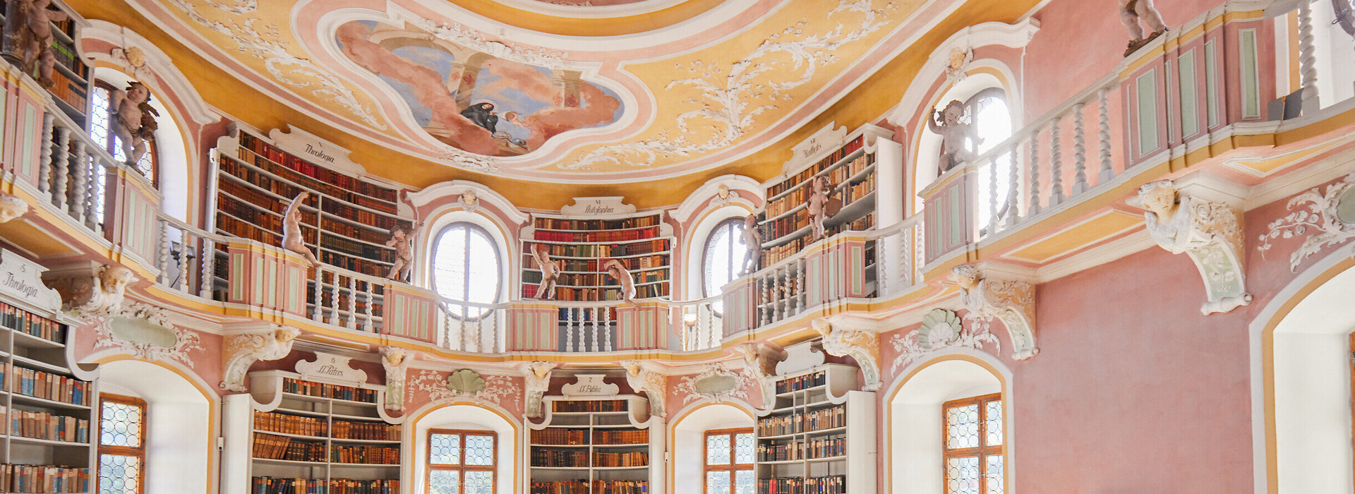 Das Bild zeigt eine imposante, kreisrunde Bibliothek mit einer hohen Decke. Die Wände sind in einem warmen, rosafarbenen Ton gemalt, und die Decke ist mit aufwendigen Mustern und wunderschönen Verzierungen geschmückt.