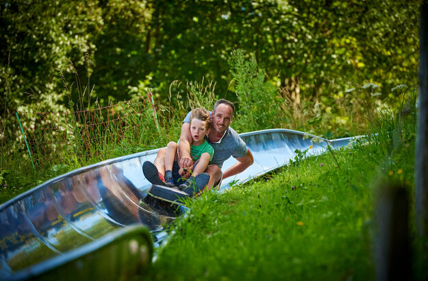 Das Bild fängt einen fröhlichen Moment eines Vaters und seines Sohnes ein, die auf einer Wasserrutsche reiten. Der Vater, der ein blaues Hemd trägt, sitzt vor dem Sohn, der ein grünes Hemd trägt. Sie halten sich beide am Rand der Rutsche fest, ihre Gesichter erhellt von Aufregung. Die Rutsche, aus blauem Plastik, ist in einem üppig grünen Feld situiert, das von Bäumen und Büschen umgeben ist. Der Vater und der Sohn sind der Hauptfokus des Bildes, ihre leuchtend bunten Hemden stehen in scharfem Kontrast zur natürlichen Kulisse. Das Bild strahlt eine Freude aus und Abenteuerlust, während sie ihre Zeit auf der Rutsche genießen.