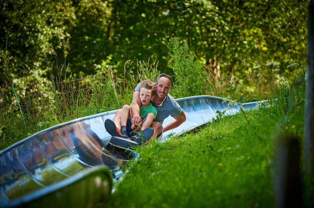 Das Bild fängt einen fröhlichen Moment eines Vaters und seines Sohnes ein, die auf einer Wasserrutsche reiten. Der Vater, der ein blaues Hemd trägt, sitzt vor dem Sohn, der ein grünes Hemd trägt. Sie halten sich beide am Rand der Rutsche fest, ihre Gesichter erhellt von Aufregung. Die Rutsche, aus blauem Plastik, ist in einem üppig grünen Feld situiert, das von Bäumen und Büschen umgeben ist. Der Vater und der Sohn sind der Hauptfokus des Bildes, ihre leuchtend bunten Hemden stehen in scharfem Kontrast zur natürlichen Kulisse. Das Bild strahlt eine Freude aus und Abenteuerlust, während sie ihre Zeit auf der Rutsche genießen.