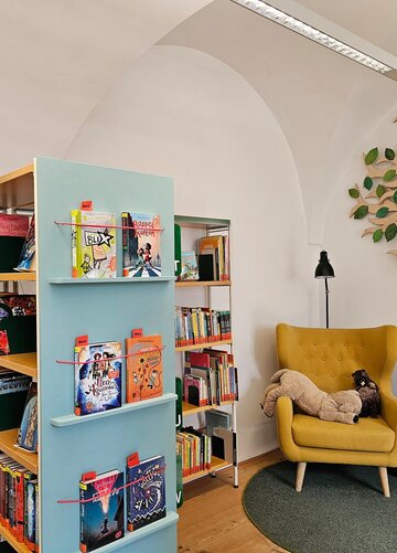 Das Bild zeigt einen Innenraum einer Bibliothek. Alles ist sehr bunt und kindgerecht dekoriert. Es ist ein mit Büchern gefülltes Regal zu sehen. Dahinter steht ein gelber sessel. An der Wand ist ein großer gebastelter Baum angebracht.