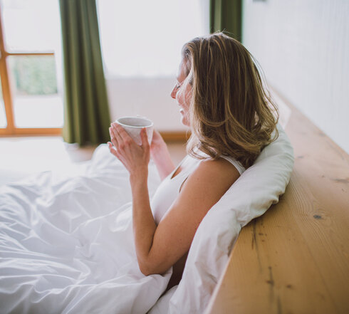 Das Bild zeigt eine Frau mit einem weißen kurzen Oberteil. Sie sitzt in einem Bett, das weiß bezogen ist. Sie lehnt an der Rückwand des Bettes und hält eine Teetasse in der Hand.