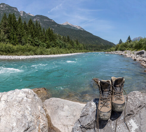 Das Bild zeigt im Vordergrund ein Paar Wanderschuhe, das auf einem felsigen Vorsprung steht. Der Fels steht am Ufer eines türkisfarbenen Flusses, des Flusses Lech. Auf dem Fluss sind leichte Stromschnellen zu sehen, was darauf hinweist, dass es sich um ein rasch fließendes Gewässer handelt. Der Fluss ist von zwei Ufern umgeben, die dicht mit Grünland bestanden sind. Im Hintergrund erheben sich majestätische Berge, deren Gipfel sich zum klaren blauen Himmel erstrecken. Die gesamte Szene suggeriert eine ruhige und idyllische Wanderdestination.