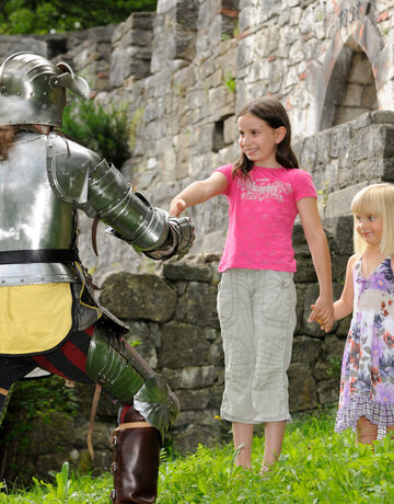 Das Bild zeigt einen Moment der Interaktion zwischen einem Ritter und zwei jungen Mädchen. Der Ritter in voller Rüstung kniet auf einem Knie in einer grünen Fläche. Er streckt seine Hand den Mädchen entgegen, die vor ihm stehen. Die Mädchen, in legerer Kleidung, halten Hände und scheinen mit dem Ritter zu sprechen. Die Kulisse des Bildes ist eine Steinmauer, die dem Bild einen historischen Kontext verleiht. Die Ritterrüstung und die Steinmauer lassen darauf schließen, dass die Szene in einem historischen oder mittelalterlichen Rahmen angesiedelt sein könnte. Die legere Kleidung der Mädchen kontrastiert mit der Rüstung des Ritters und hebt den Unterschied ihrer Rollen und der Zeitperioden, die sie repräsentieren, hervor. Das Bild enthält keine Texte.
