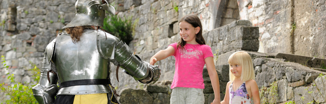 Das Bild zeigt einen Moment der Interaktion zwischen einem Ritter und zwei jungen Mädchen. Der Ritter in voller Rüstung kniet auf einem Knie in einer grünen Fläche. Er streckt seine Hand den Mädchen entgegen, die vor ihm stehen. Die Mädchen, in legerer Kleidung, halten Hände und scheinen mit dem Ritter zu sprechen. Die Kulisse des Bildes ist eine Steinmauer, die dem Bild einen historischen Kontext verleiht. Die Ritterrüstung und die Steinmauer lassen darauf schließen, dass die Szene in einem historischen oder mittelalterlichen Rahmen angesiedelt sein könnte. Die legere Kleidung der Mädchen kontrastiert mit der Rüstung des Ritters und hebt den Unterschied ihrer Rollen und der Zeitperioden, die sie repräsentieren, hervor. Das Bild enthält keine Texte.
